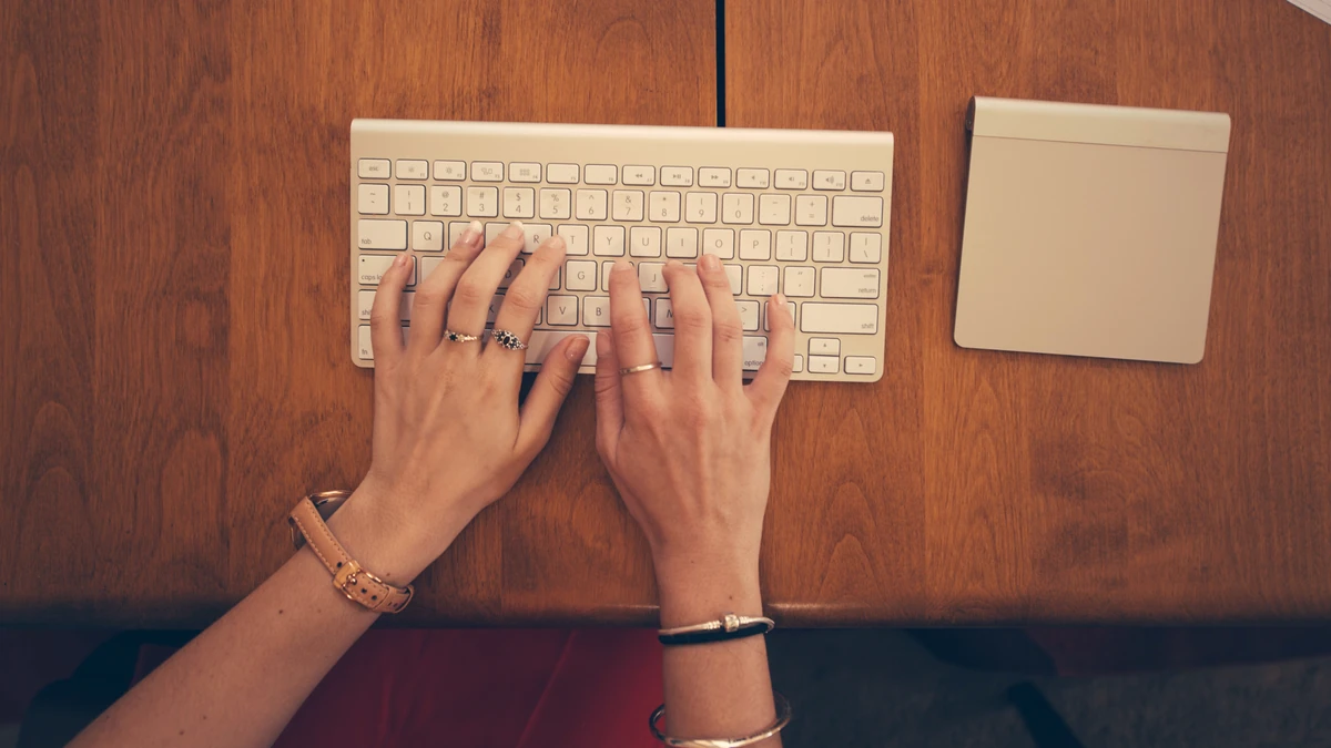 Two hands typing on a white wireless keyboard on a wooden desk