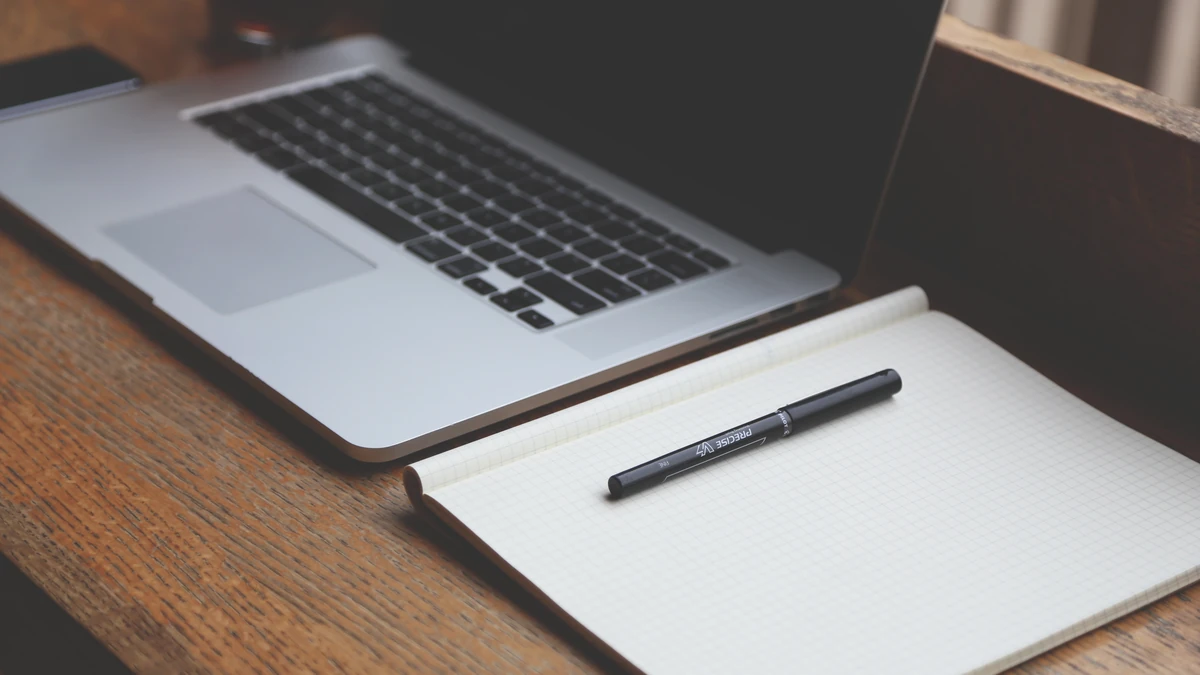Laptop beside an open lined notebook with a black pen on a wooden desk